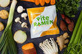 A pack of Vite Ramen Vegan Japanese Curry v3.0 on a stone countertop surrounded by fresh ingredients such as onion, leek, potato, carrot, celery, ginger root, and herbs.